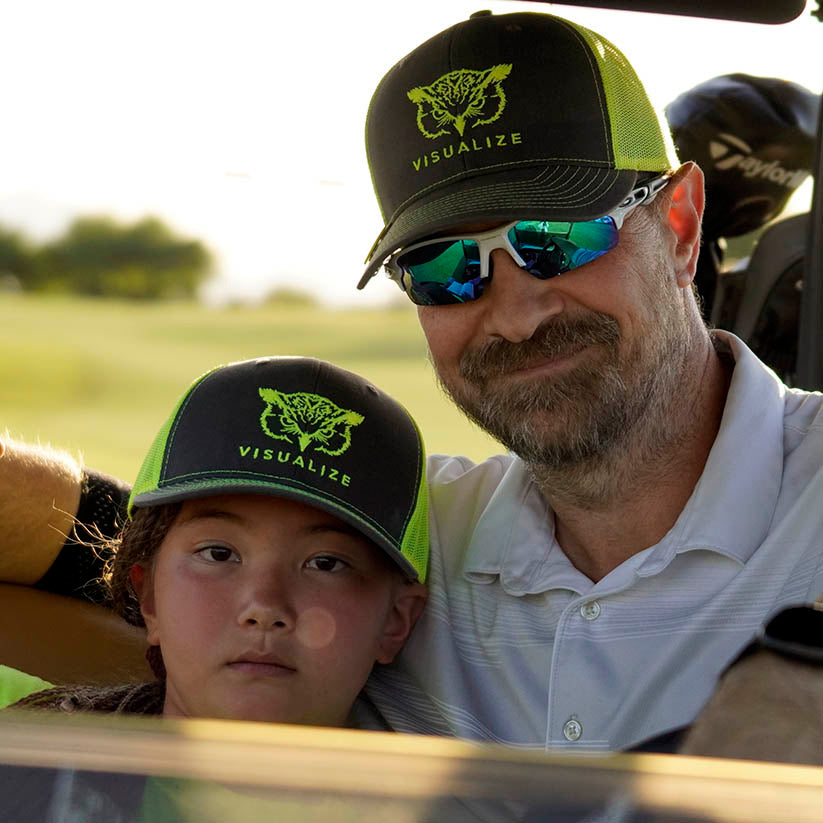 A young golfer with her dad wearing awesome yellow and gray visualize hats. 